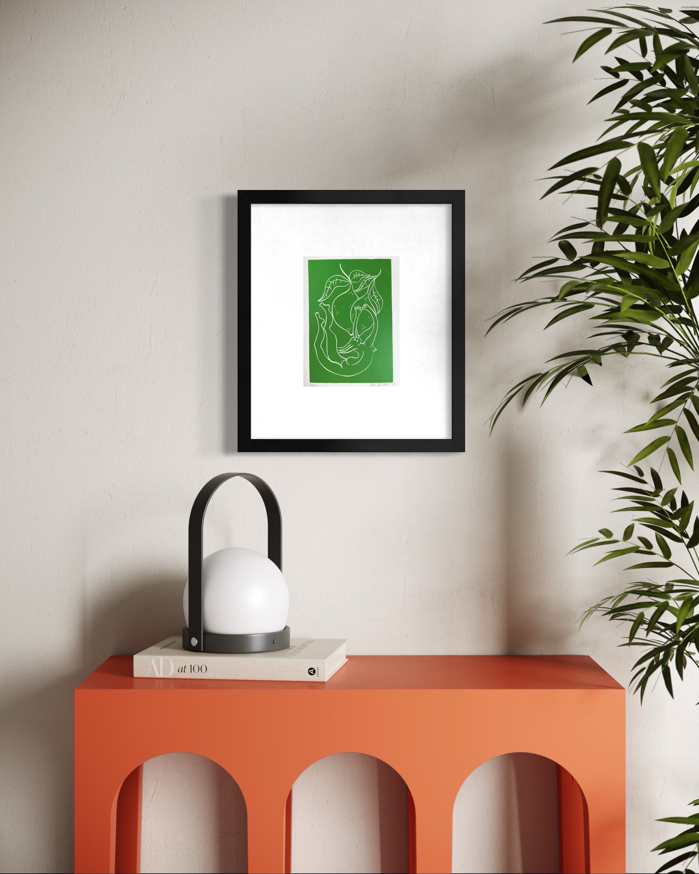 Framed green artwork on a wall above an orange table with a lamp and books.