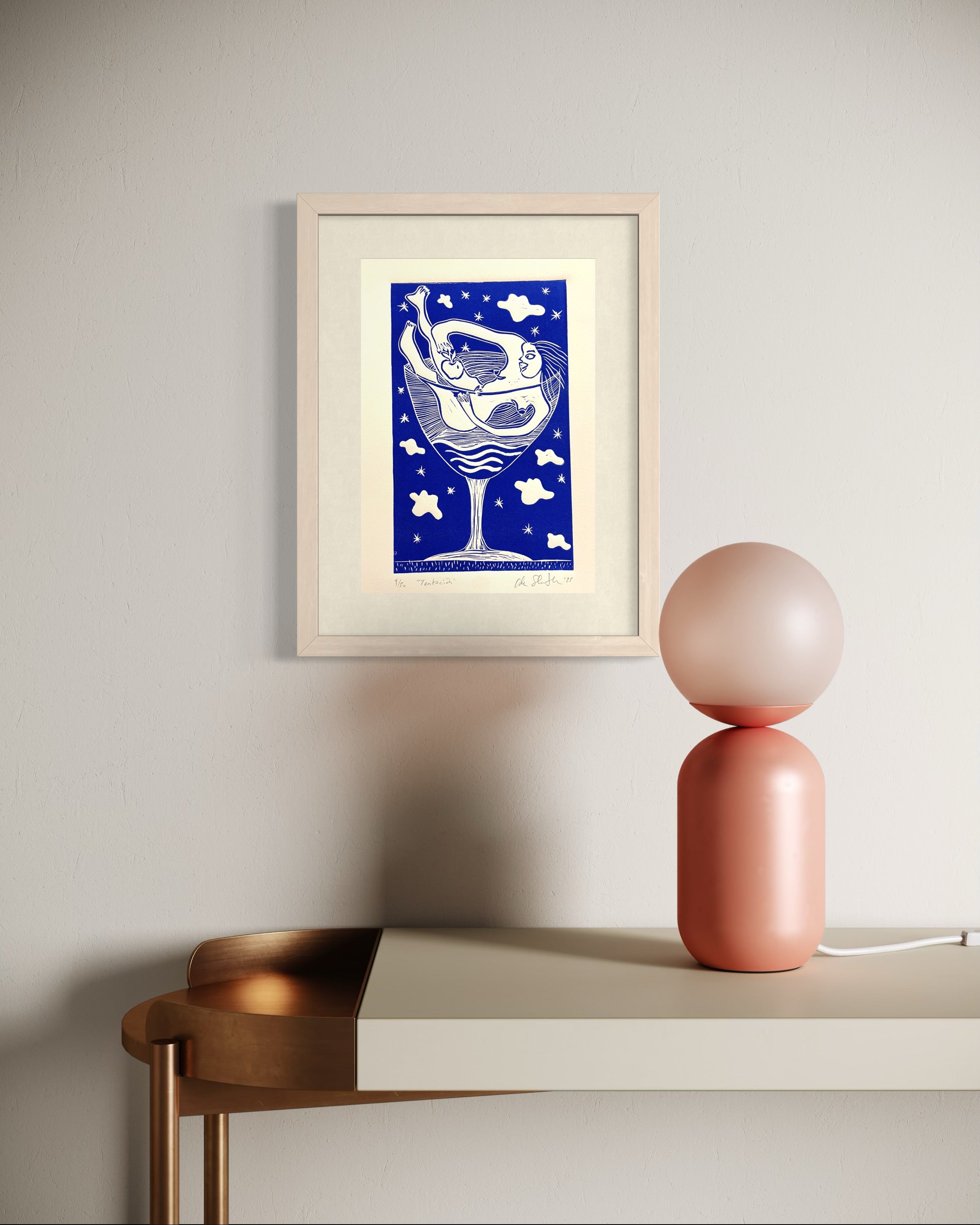 Framed artwork on a wall above a table with a pink lamp.