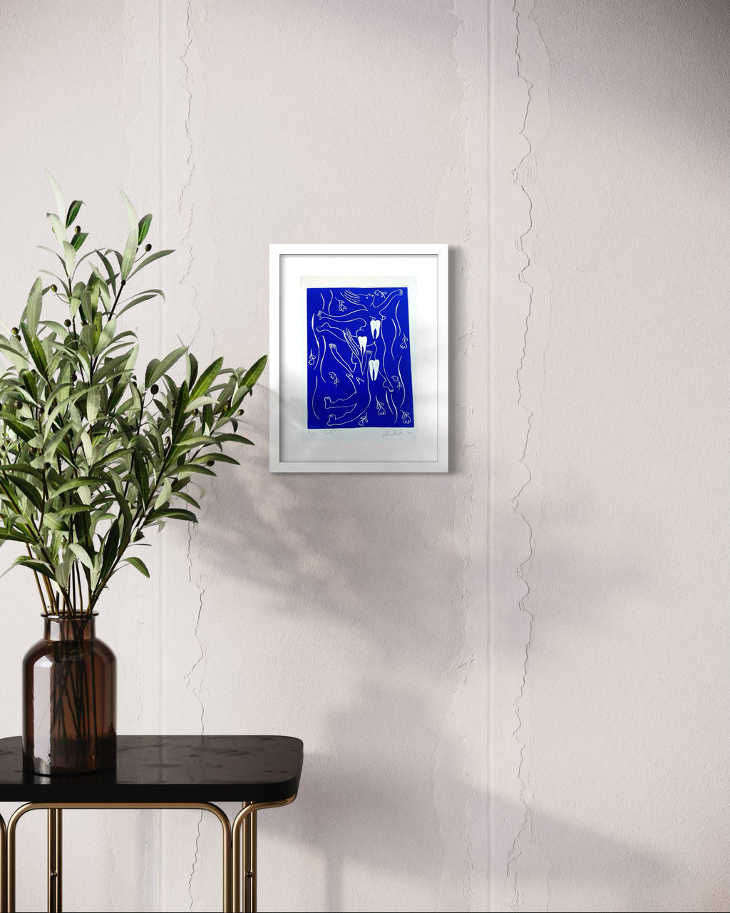 Framed abstract blue artwork on a white wall with a vase of green leaves on a black table.