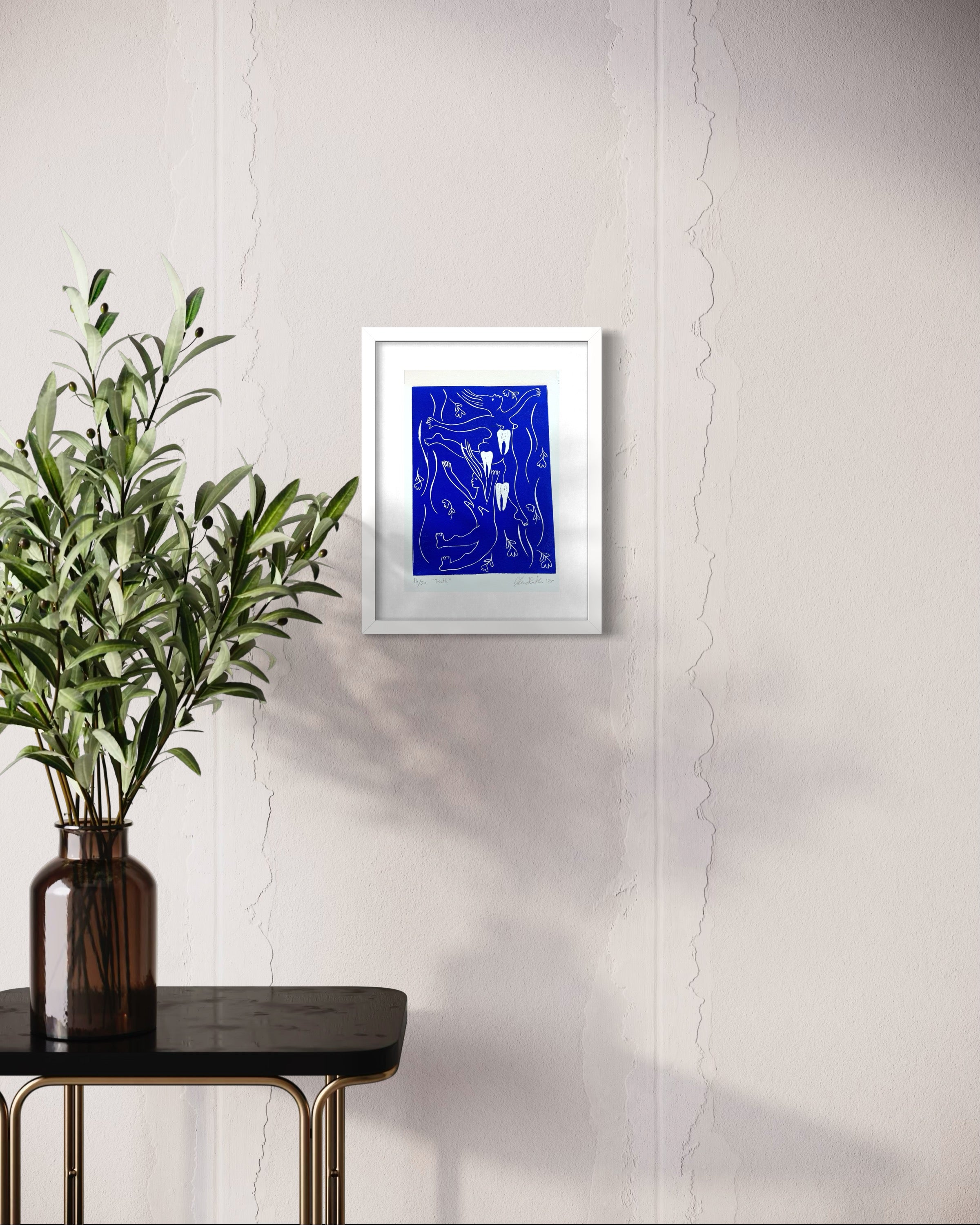 Framed abstract blue artwork on a white wall with a vase of green leaves on a black table.