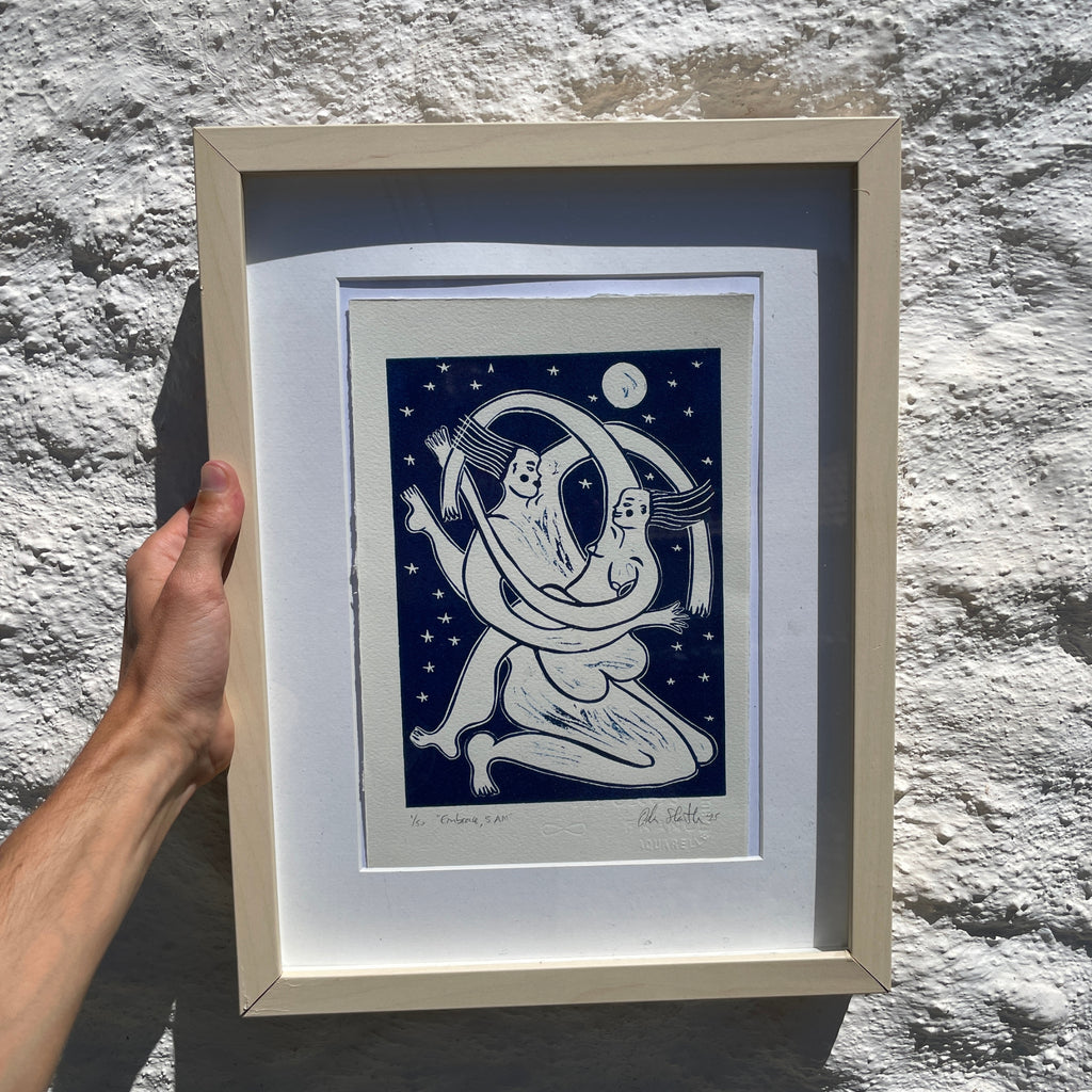 Framed artwork of two people embracing under a crescent moon against a starry night sky, held by a hand against a textured stone wall.