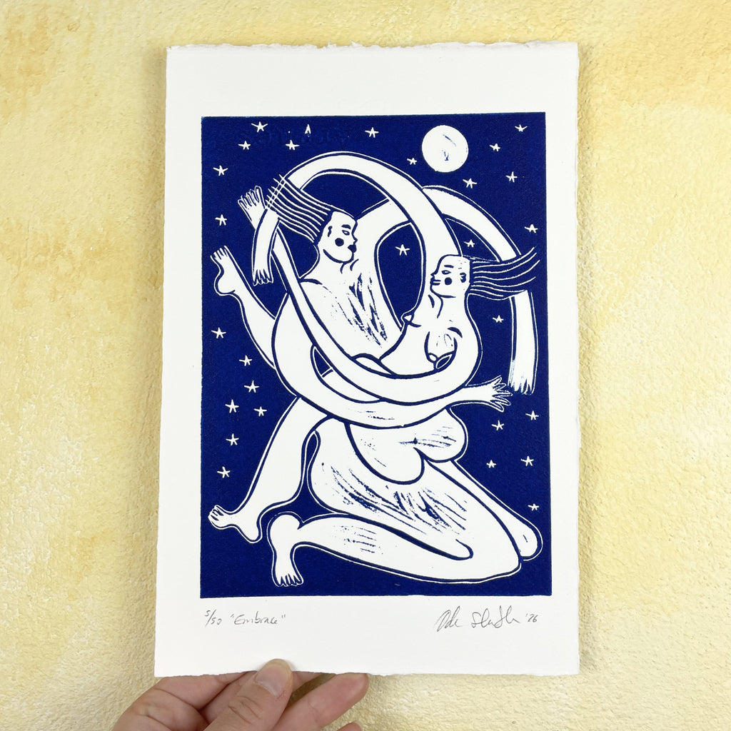 Art print of two abstract figures embracing on a blue background with stars and a moon, held against a beige wall.