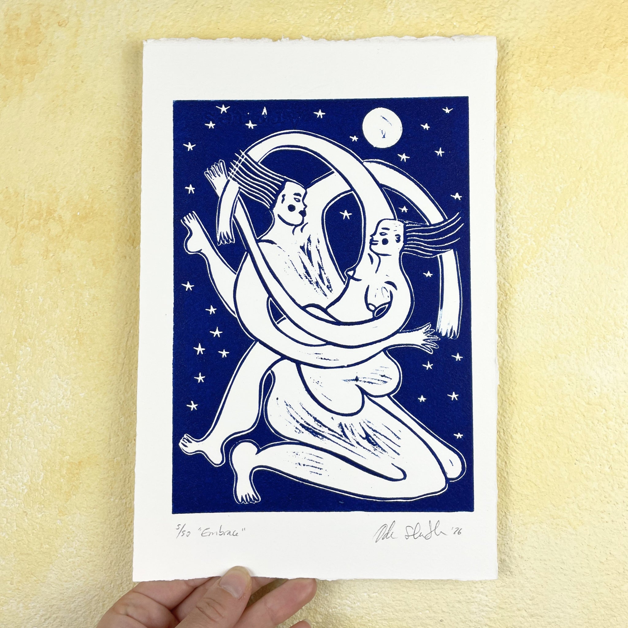 Art print of two abstract figures embracing on a blue background with stars and a moon, held against a beige wall.
