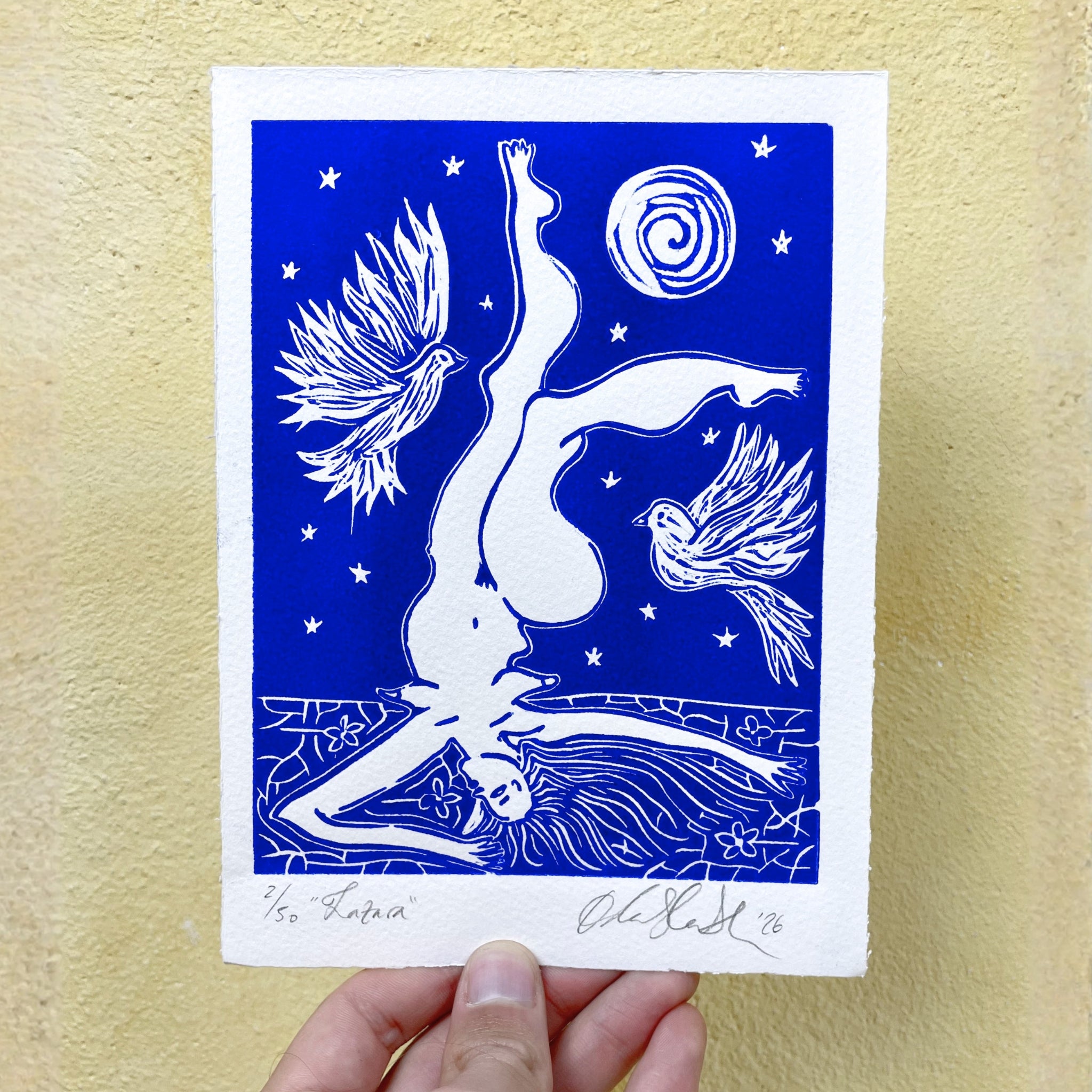 Art print with a white figure and birds on a blue background, held against a yellow wall.