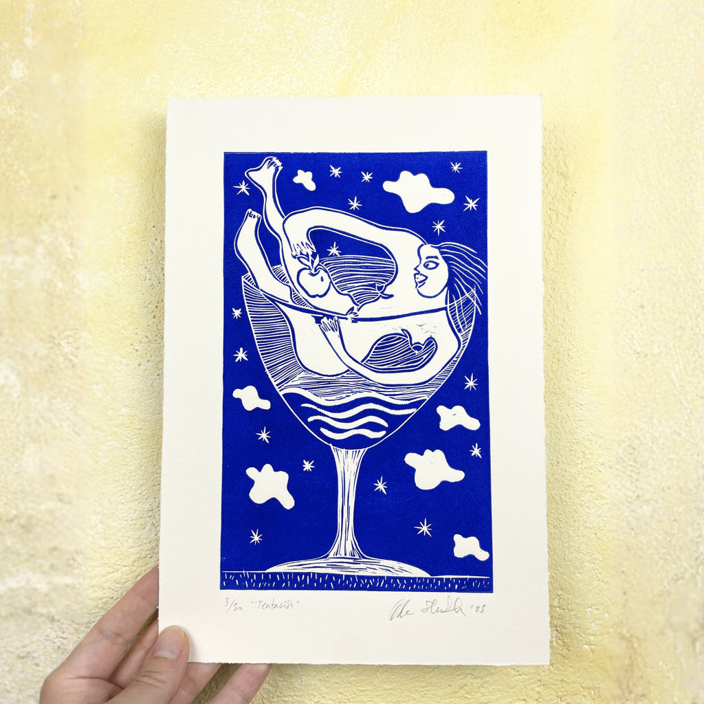 Playful blue linocut "Tentación", a print of a woman relaxing in a martini glass holding an apple with abstract figures and clouds on a blue background, held against a textured yellow wall.