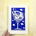 Playful blue linocut "Tentación", a print of a woman relaxing in a martini glass holding an apple with abstract figures and clouds on a blue background, held against a textured yellow wall.