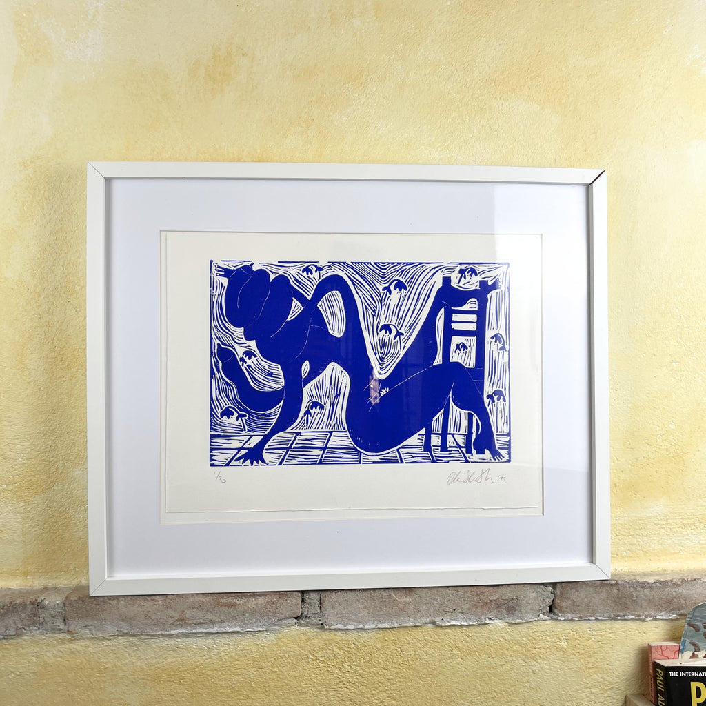 Framed artwork of a blue abstract design on a yellow wall. Ultramarine blue figurative linocut "Twisted" of a nude body turning in a room with a chair